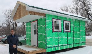 A man stands beside a tiny home. There is snow on the ground and the sky is cloudy.