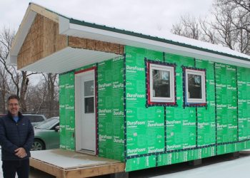 A man stands beside a tiny home. There is snow on the ground and the sky is cloudy.