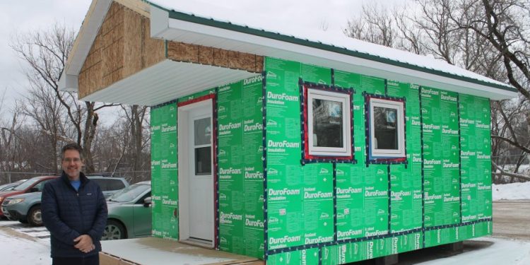 A man stands beside a tiny home. There is snow on the ground and the sky is cloudy.