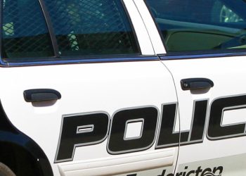 A close-up photo shows the passenger side of a white and black or dark blue police car. The word POLICE is written in dark letters across the doors and a Canadian flag decal is also visible in the upper left-hand corner of the shot.