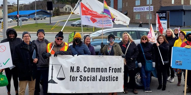 A crowd stands holding protest signs and a banner with "NB Common Front for Social Justice" written on it.
