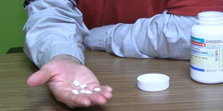 Several white pills are held in the palm of someone's hand. A pill bottle sits on the surface of a table.