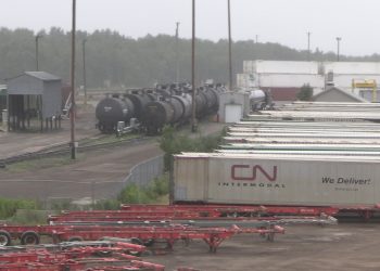 Shipping containers and other equipment sit idle in a yard beside a railway.